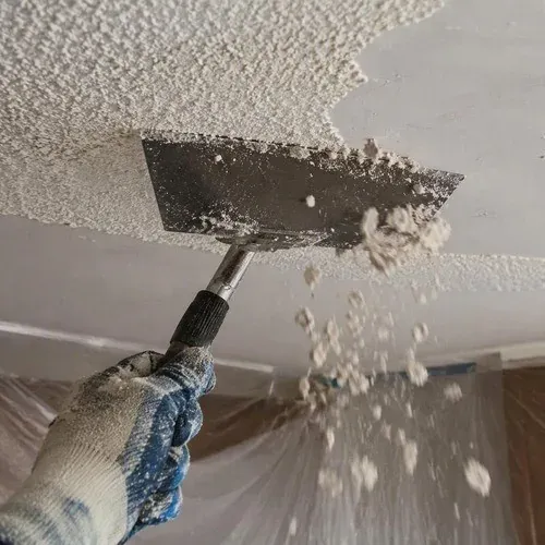 Scraping wetted popcorn texture off a bedroom ceiling, which keeps dust down and protects the board
